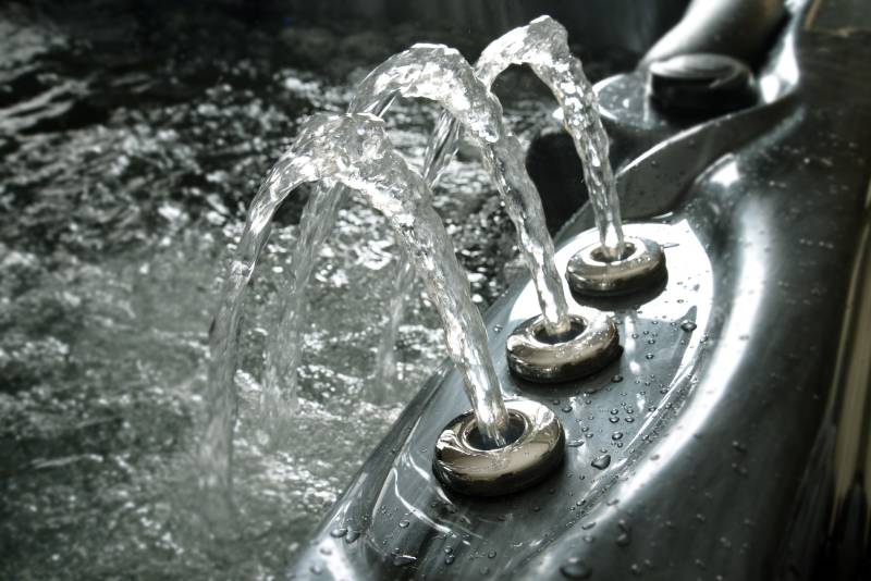 Trois jets d'eau clairs jaillissent d'un spa moderne noir, créant des arcs scintillants au-dessus de l'eau agitée. Détente et luxe.