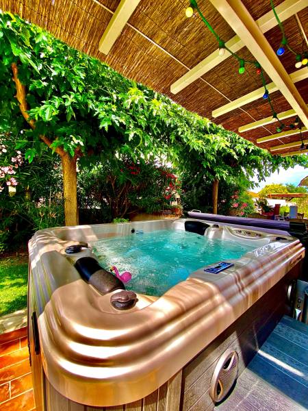 Jacuzzi extérieur bouillonnant sous une pergola en paille et feuillage luxuriant. Guirlandes lumineuses colorées créent une ambiance détente.