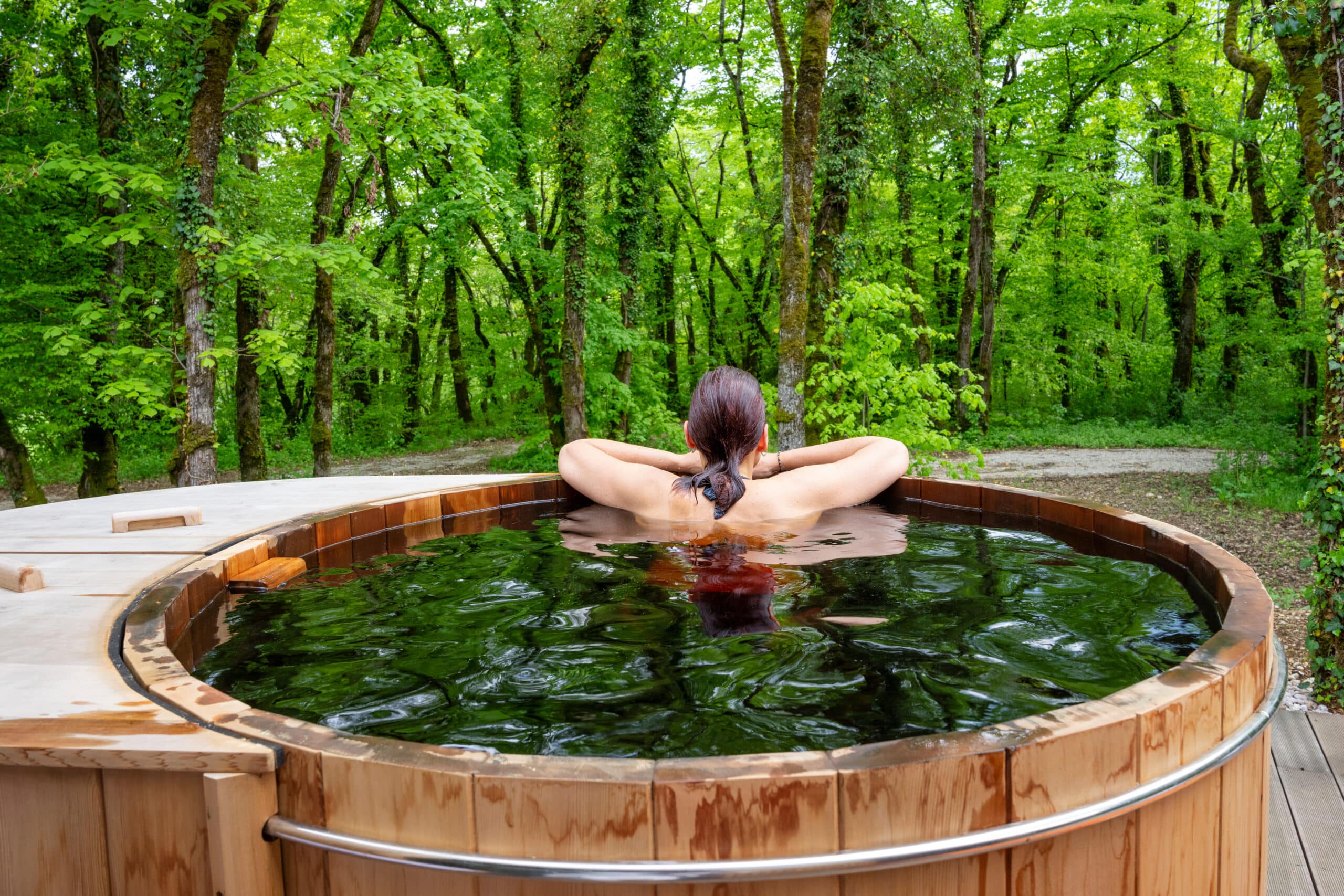 Femme détendue dans un bain nordique en bois, dos tourné vers une forêt verdoyante luxuriante. L'eau du spa reflète les arbres.