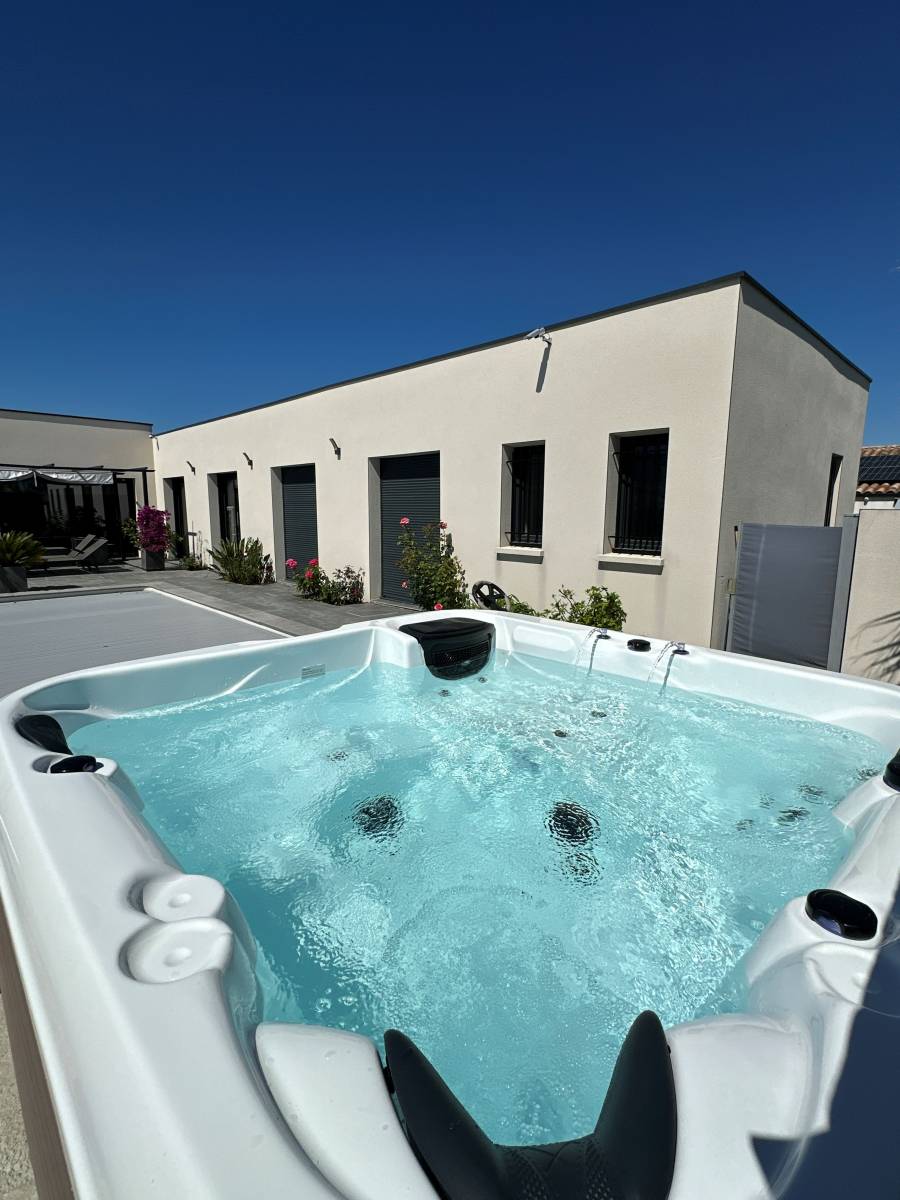 Jacuzzi moderne bouillonnant sur une terrasse ensoleillée devant une maison contemporaine beige sous un ciel bleu clair.