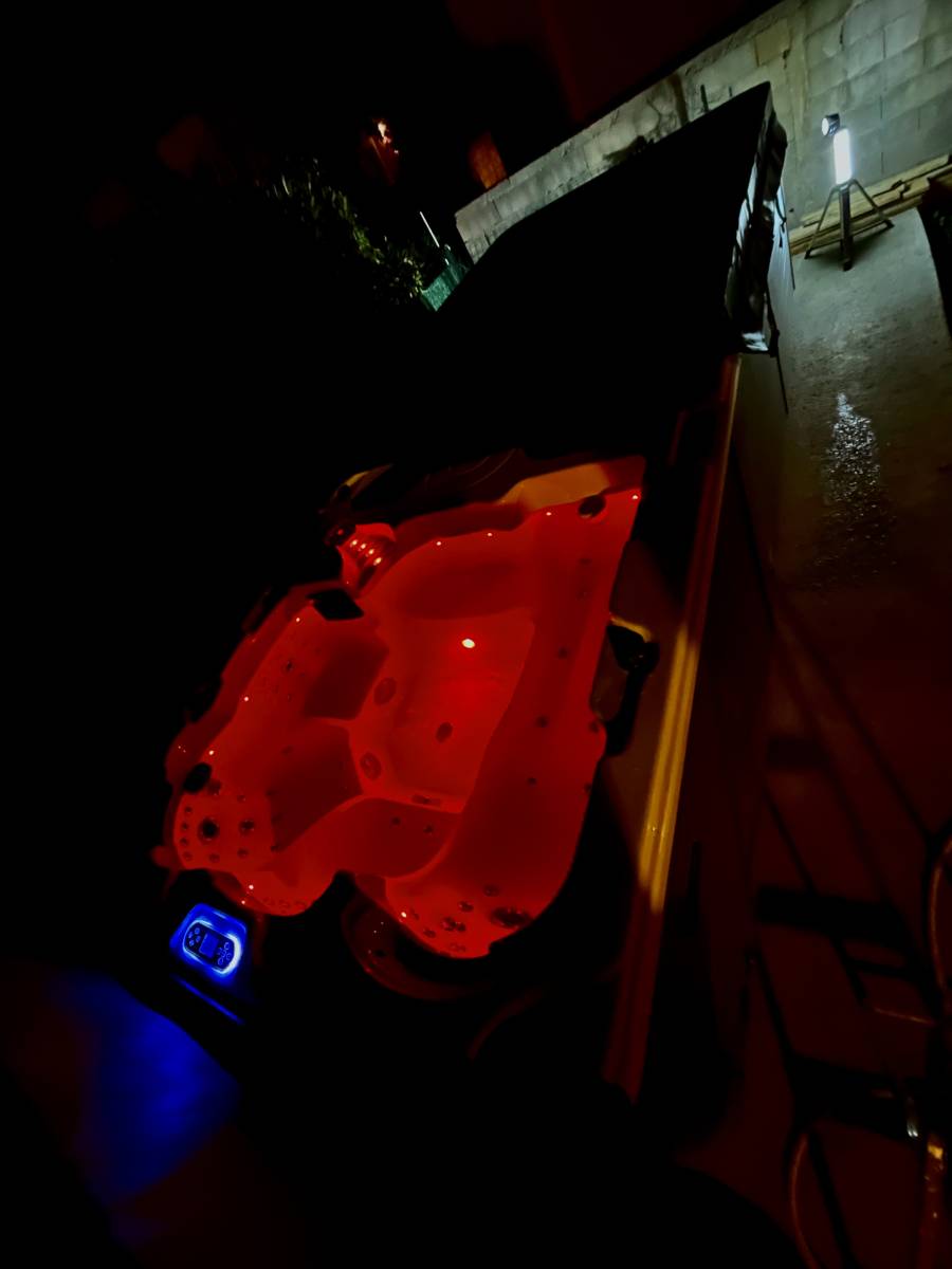 Jacuzzi extérieur éclairé en rouge vif la nuit, créant une ambiance relaxante. Panneau de contrôle bleu lumineux visible.