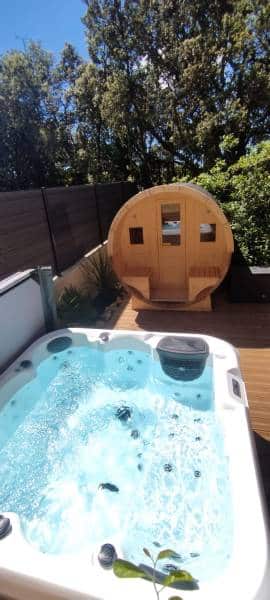 Jacuzzi bouillonnant sur une terrasse en bois, avec un sauna tonneau en bois clair à l'arrière-plan, sous un ciel bleu. Ambiance détente extérieure.