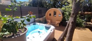 Jardin de détente : jacuzzi bleu bouillonnant et sauna tonneau en bois sur une terrasse en bois. Végétation luxuriante, arbre et clôture discrète.