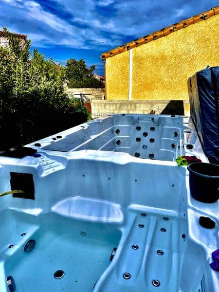 Deux jacuzzis blancs vides en extérieur, dont un nettoyé et humide. Arbres verts, bâtiment jaune, ciel bleu nuageux. Préparation avant remplissage.