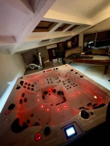 Jacuzzi éclairé rouge dans salle de loisirs sous combles. Billard, elliptique et plafond mansardé visible. Ambiance détente moderne.
