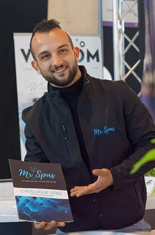 Homme souriant, barbu, en veste "Mr Spas", présente un "CATALOGUE SPAS" pour le bien-être. Geste accueillant.