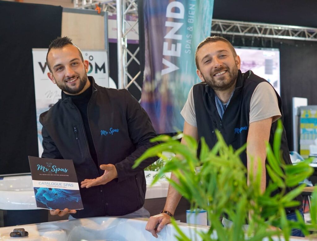 Hommes souriants présentant le catalogue « Mr Spas » et un spa. L'un tient la brochure. Banderole « SPAS & BIEN ÊTRE » visible.