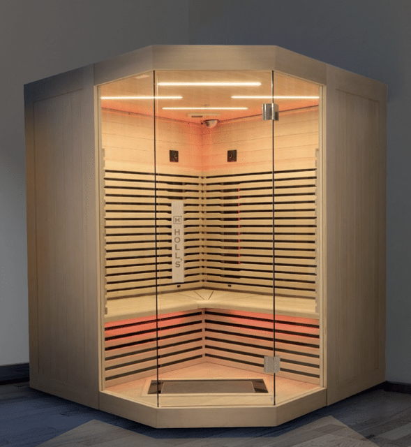 Sauna d'angle moderne en bois clair avec porte vitrée, éclairage LED blanc au plafond et rouge sous les bancs. Le panneau vertical à l'intérieur affiche le texte 'HOLLS'.