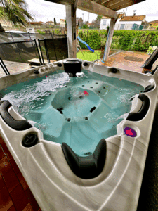 Spa extérieur moderne avec eau bouillonnante et lumières colorées, sous une pergola, sur une terrasse avec jardin en arrière-plan.