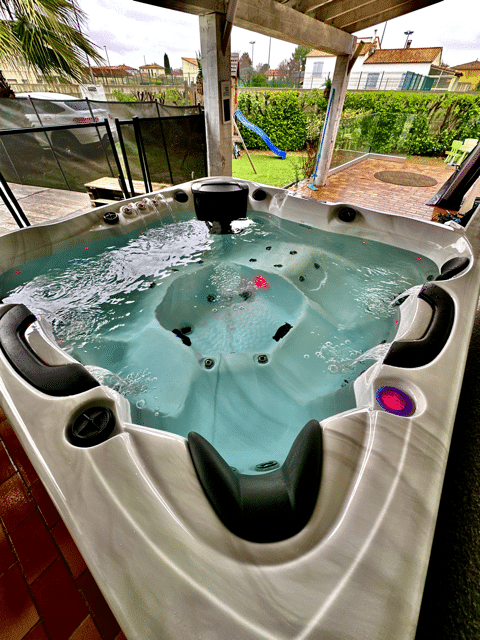 Spa extérieur moderne avec eau bouillonnante et lumières colorées, sous une pergola, sur une terrasse avec jardin en arrière-plan.