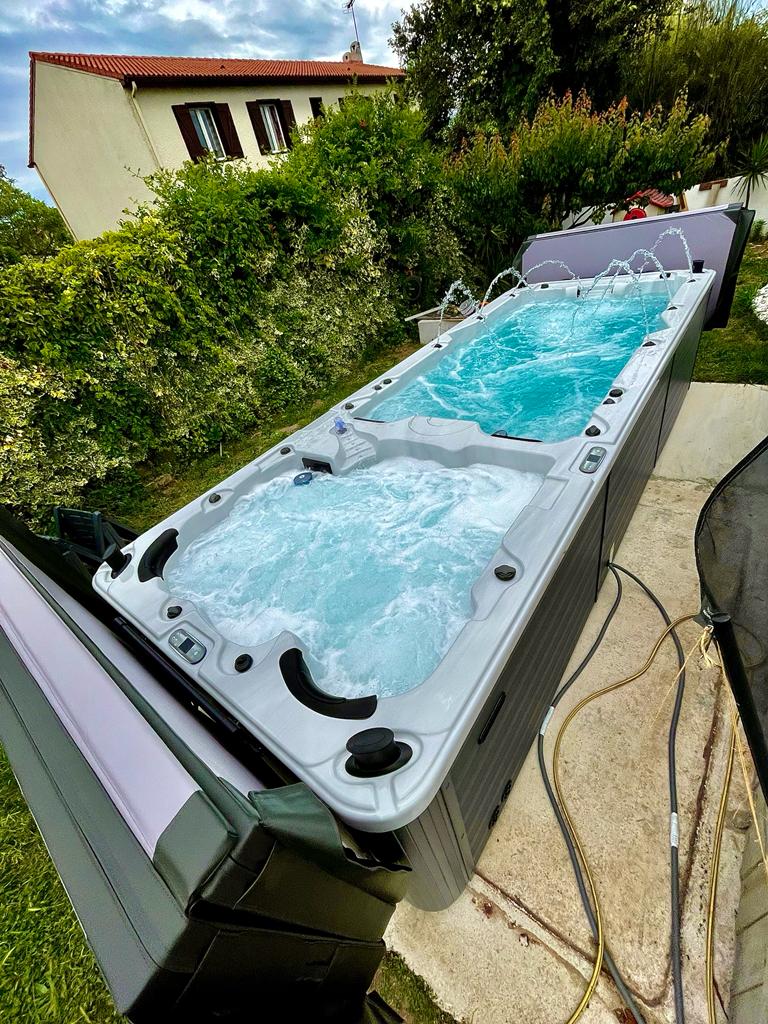 Spa de nage extérieur avec jacuzzi bouillonnant et jets d'eau, installé près d'une maison avec toit rouge et végétation luxuriante.