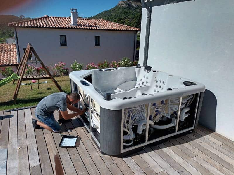 Technicien agenouillé réparant un jacuzzi sur une terrasse en bois. Les panneaux du spa sont ouverts, révélant les tuyaux et pompes.