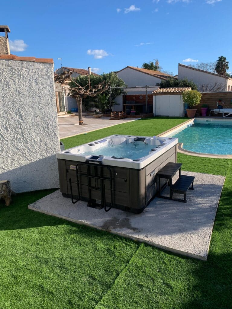Luxueux spa de jardin bouillonnant (jacuzzi) près d'une piscine et d'une terrasse ensoleillée. Gazon synthétique vert et ciel bleu clair.