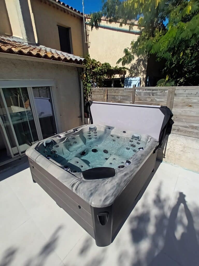 Spa extérieur moderne avec couverture ouverte, bulles actives, installé sur une terrasse blanche près d'une maison et d'une clôture en bois sous un ciel bleu.