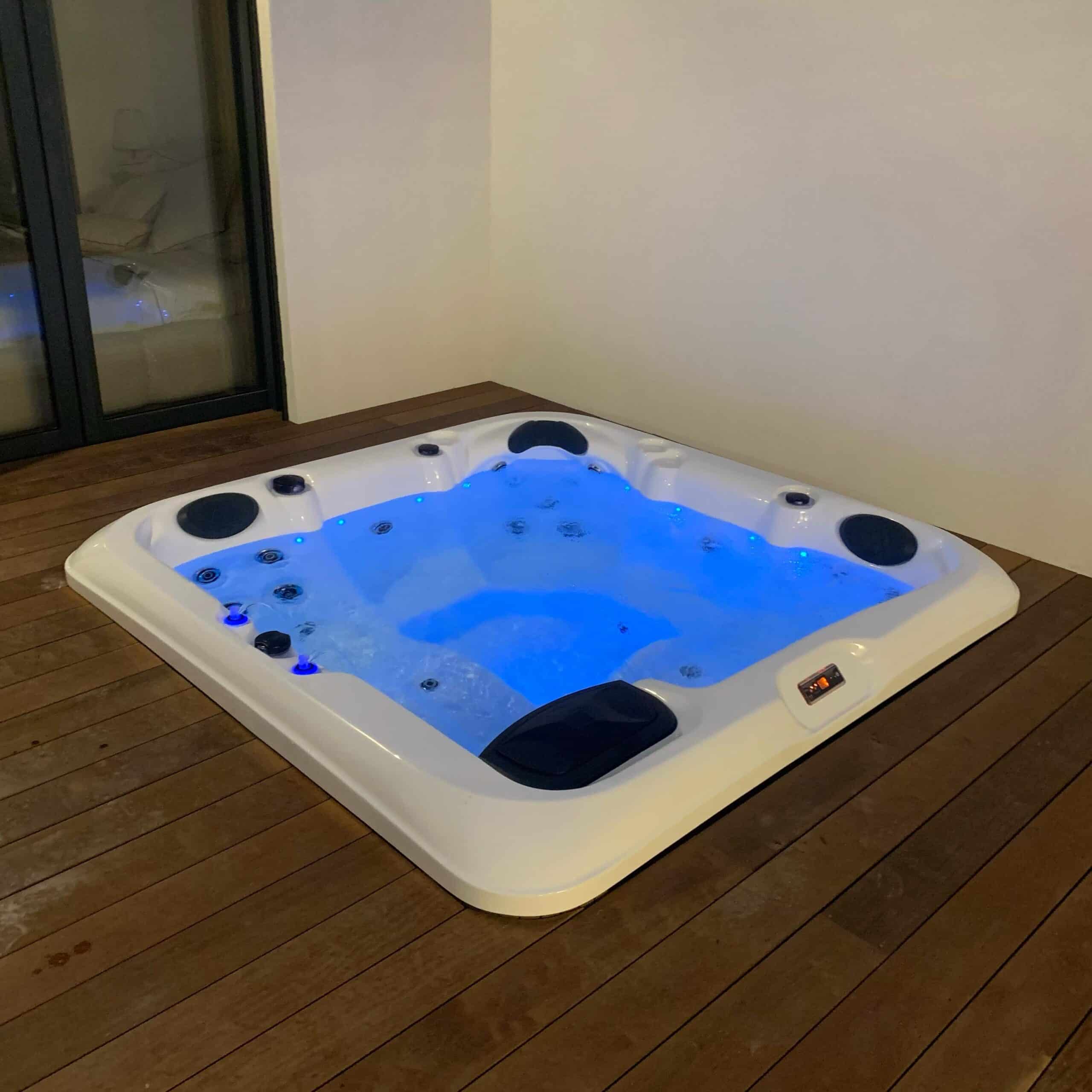 Spa gonflable carré blanc éclairé par des LED bleues, en activité sur une terrasse en bois sombre près d'une baie vitrée.