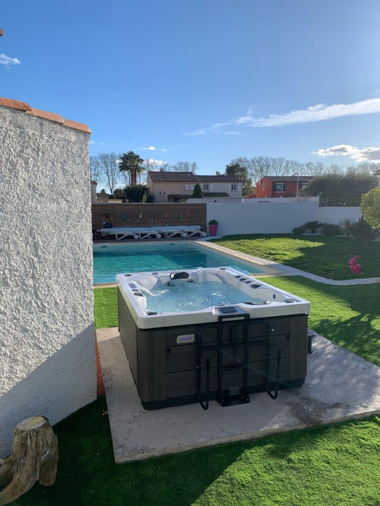 Spa Vendom : Profitez du jacuzzi extérieur près de la piscine. Spa jacuzzi Vendom en fonctionnement dans un jardin ensoleillé. Vue sur la piscine turquoise, la pelouse artificielle et le ciel bleu.